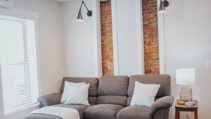 A living room with a gray sofa, two white pillows, a small wooden side table with a lamp, and a brick accent wall. - Charming 2-bedroom apartment with AC, WiFi in lovely Mifflinburg (Mifflinburg)