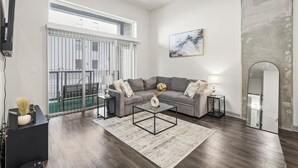 A modern living room with a grey sofa, a glass coffee table, and a large mirror. There is a sliding glass door leading to a balcony.