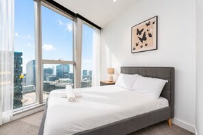 Ein Hotelzimmer mit großem Fenster, einem Bett, einem Nachttisch und Blick auf die Stadt.