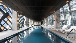 An indoor swimming pool with clear glass walls, lounge chairs, and a modern architectural design.