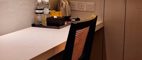 A hotel room with a desk, a chair, a kettle, water bottles, and a tissue box.