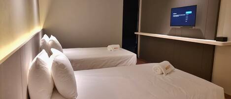 A hotel room with two single beds, each with a pillow and a towel with the name 'ROXY'. A flat-screen TV is mounted on the wall above a shelf.