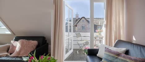 Una sala de estar moderna con un sofá de cuero negro, un sillón negro y un sillón blanco. Hay un ventanal con vistas a un edificio de piedra y un balcón con mesa y sillas.