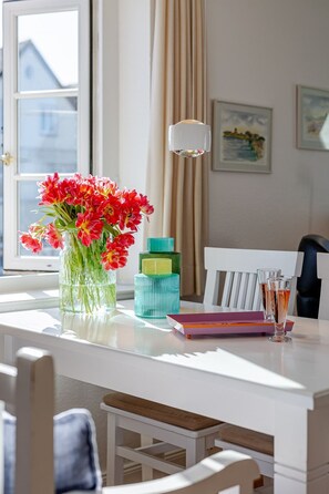 A dining area with a white table, a vase with red flowers, a green bottle, and a white pendant light.