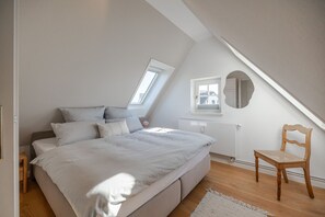 A bedroom with a sloped ceiling, a bed with white bedding, a wooden chair, and a small window.