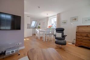 Ein modernes Wohnzimmer mit Flachbildfernseher, Couchtisch aus Holz, schwarzem Ledersessel, Holzschrank und Essbereich mit weißen Stühlen und Tisch.
