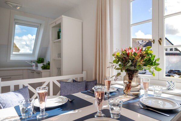 Un comedor con una mesa dispuesta para dos, un florero con flores y vistas al cielo a través de la ventana.