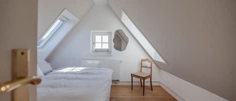 A bedroom with a sloped ceiling, a bed, a chair, and a window.