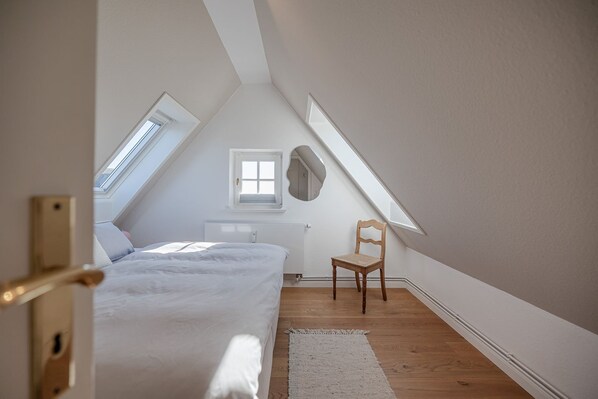 A bedroom with a sloped ceiling, a bed, a chair, and a window.