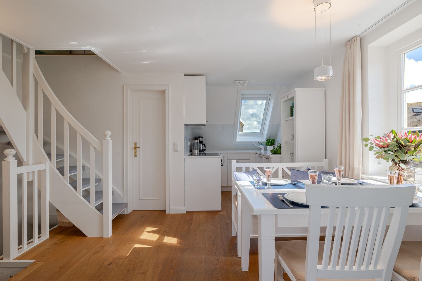 Eine moderne Küche mit weißen Schränken, ein Essbereich mit einem Tisch für zwei Personen und eine Treppe mit weißen Geländern.