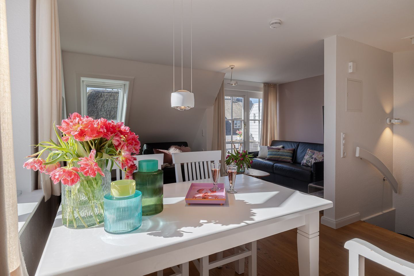 A dining area with a white table, chairs, and a vase of pink flowers. There is a window with curtains, a sofa in the background, and a hanging light fixture.