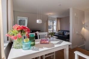 A dining area with a white table, chairs, and a vase of pink flowers. There is a window with curtains, a sofa in the background, and a hanging light fixture.