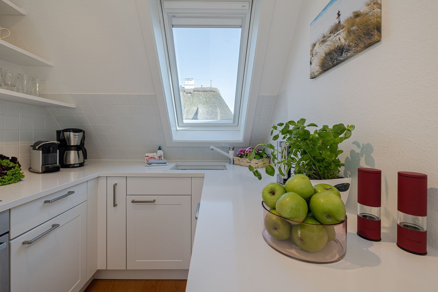 Une cuisine moderne avec des meubles blancs, une machine à café, un saladier de pommes vertes et une plante en pot.