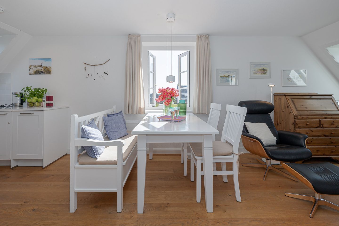 Un espace de repas moderne et bien éclairé, avec une table blanche, des chaises blanches et un fauteuil en cuir noir. On y trouve une fenêtre avec des rideaux, un meuble blanc orné d’une plante, et des œuvres d’art accrochées aux murs.