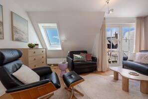 Una sala moderna con un sofá de cuero negro, una mesa de centro de madera y un sillón negro. Hay una ventana con vistas al cielo y una puerta que da a un balcón.