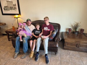Family relaxes on a comfortable couch in a cozy room with a framed photograph, table lamp, and potted plant.