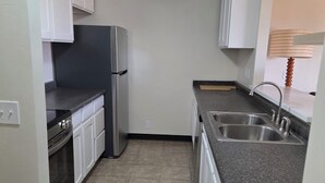 A compact kitchen with white cabinets, a stainless steel refrigerator, a built-in oven, a sink with a faucet, and a ceiling light. - New Western Inn & Suites (Fillmore)