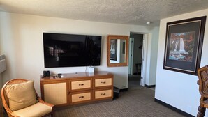 A hotel room with a large flat-screen TV, a wooden dresser with wicker drawers, a wicker chair, a framed picture on the wall, and a carpeted floor. - New Western Inn & Suites (Fillmore)