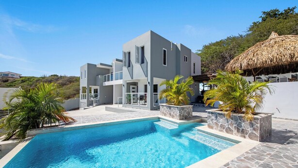 Une maison moderne à deux étages, dotée d’une piscine, de jardinières en pierre et d’une structure à toit de chaume.