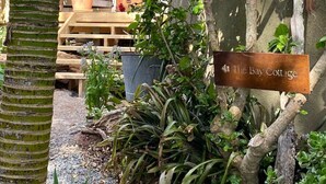 Un'area esterna rustica con una panca di legno, un cartello con su scritto 'The Bay Cottage', e diverse piante e alberi.