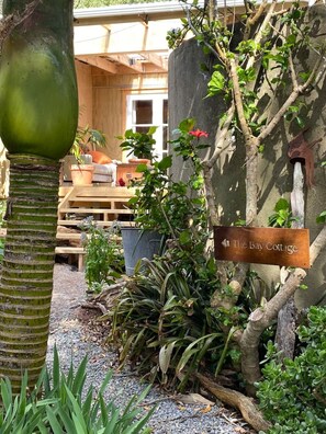 Ein rustikaler Außenbereich mit einer Holzbank, einem Schild mit der Aufschrift 'The Bay Cottage' und verschiedenen Pflanzen und Bäumen.