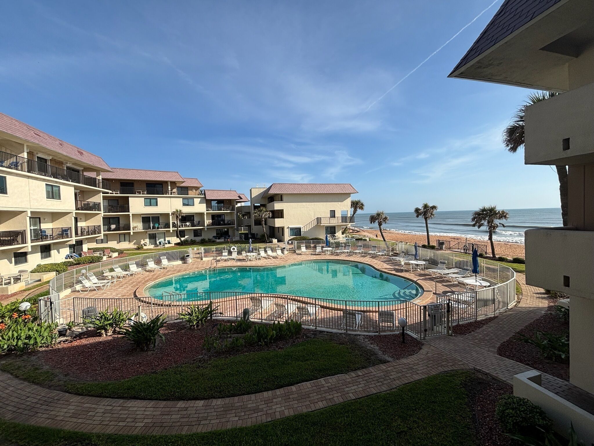 A resort with a swimming pool surrounded by lounge chairs and palm trees, overlooking a beach.