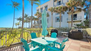 Une terrasse extérieure avec des chaises turquoise et une table, entourée de palmiers et d’un immeuble doté de balcons.