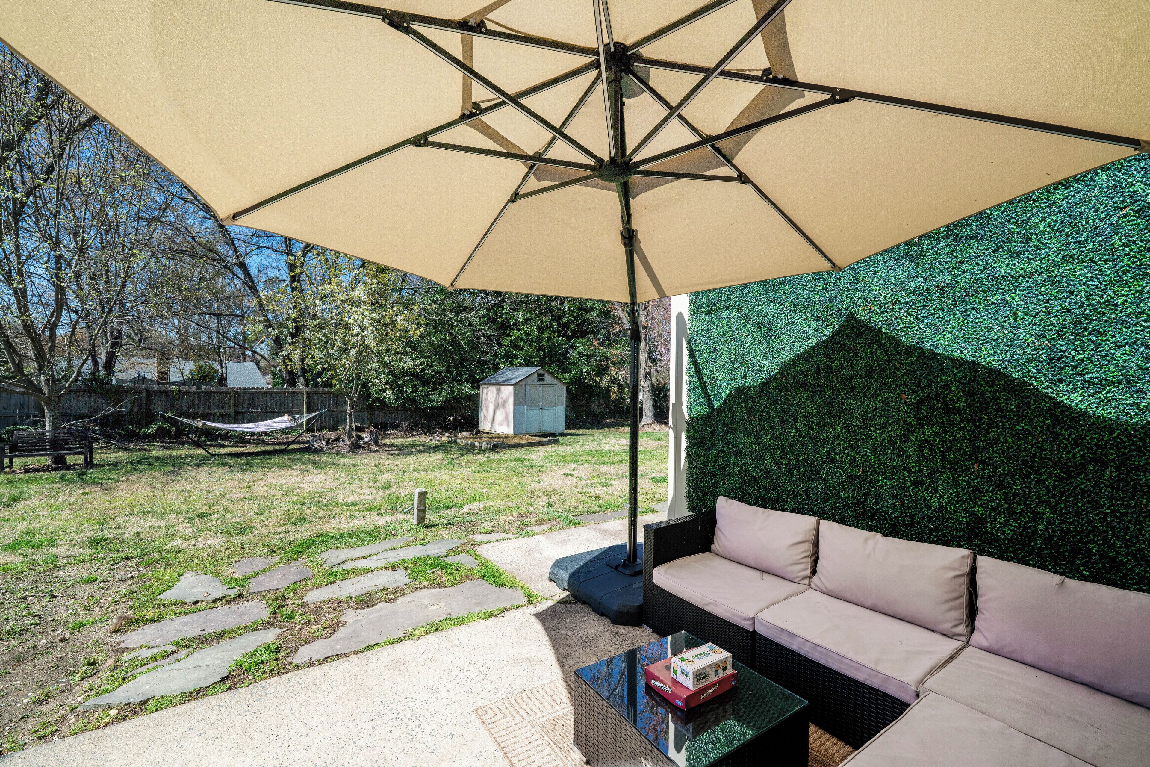 Un área de patio con un paraguas color beige, un espacio de asientos con cojines y un muro de seto artificial verde.
