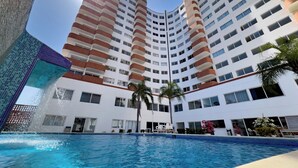 Un hotel moderno con piscina, varios balcones y un cielo azul despejado.