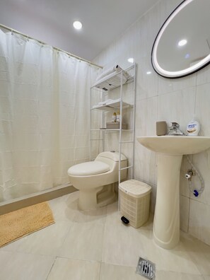 Casa de banho com sanita, lavatório, espelho, prateleira com produtos de higiene pessoal e cortina de chuveiro.