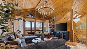 A cozy living room with a large sofa, a coffee table, and a fireplace. There is a TV mounted on the wall and a chandelier hanging from the ceiling. - Spring Break for families in BR with Sauna, Hot tub, Firepit & amazing views (Blue Ridge)