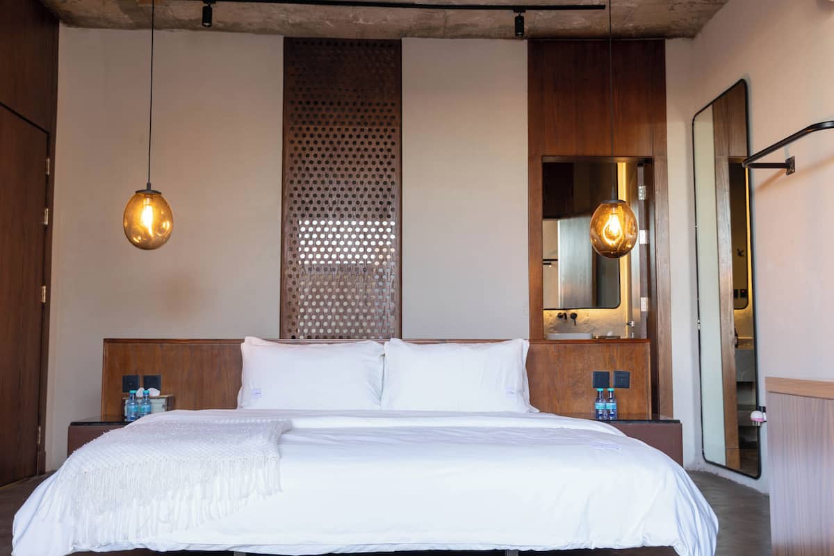 Una cama bien hecha con ropa blanca, dos mesitas de noche con botellas, un cabecero de madera, un espejo grande y una lámpara colgante.