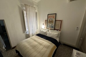 Une chambre à coucher avec un lit, une fenêtre avec des rideaux, un certificat encadré accroché au mur et un radiateur.
