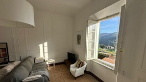 Ein Wohnzimmer mit Sofa, Sessel, Fernseher und einem Fenster mit Blick auf eine Landschaft.