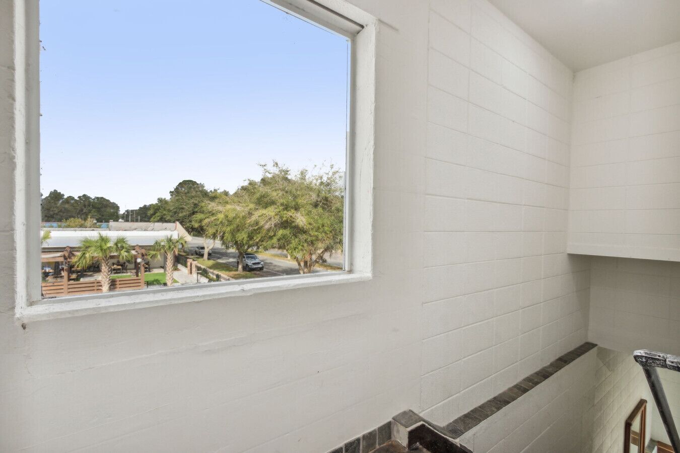 Una habitación con paredes blancas, ventana que da a un estacionamiento con árboles y un edificio.