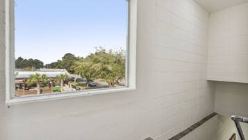 Una habitación con paredes blancas, ventana que da a un estacionamiento con árboles y un edificio.