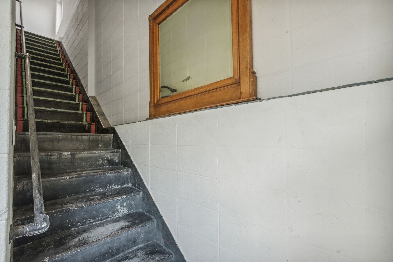 Een trap met een leuning, leidend naar een ruimte met witte betegelde muren en een raam met houten kozijn.