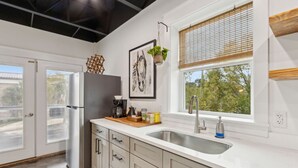 Eine moderne Küche mit weißer Arbeitsplatte, Spüle aus Edelstahl und einem Fenster mit Jalousien. Ein Kühlschrank, eine Kaffeemaschine und eine hängende Pflanze sind ebenfalls vorhanden.