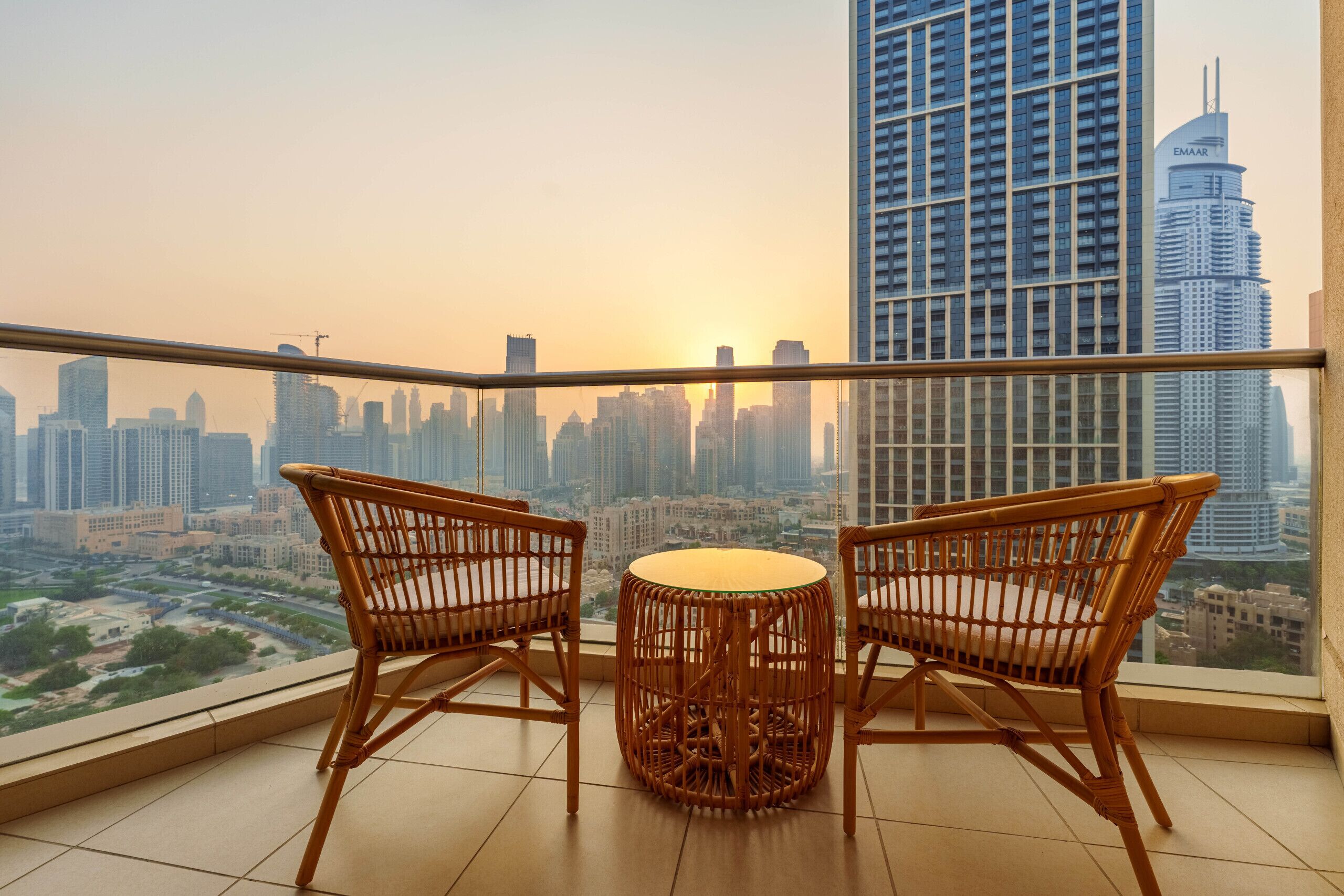 Un balcon agrémenté de deux fauteuils en osier et d’une petite table ronde, offrant une vue sur le panorama urbain, avec ses gratte-ciel et son ciel dégagé.