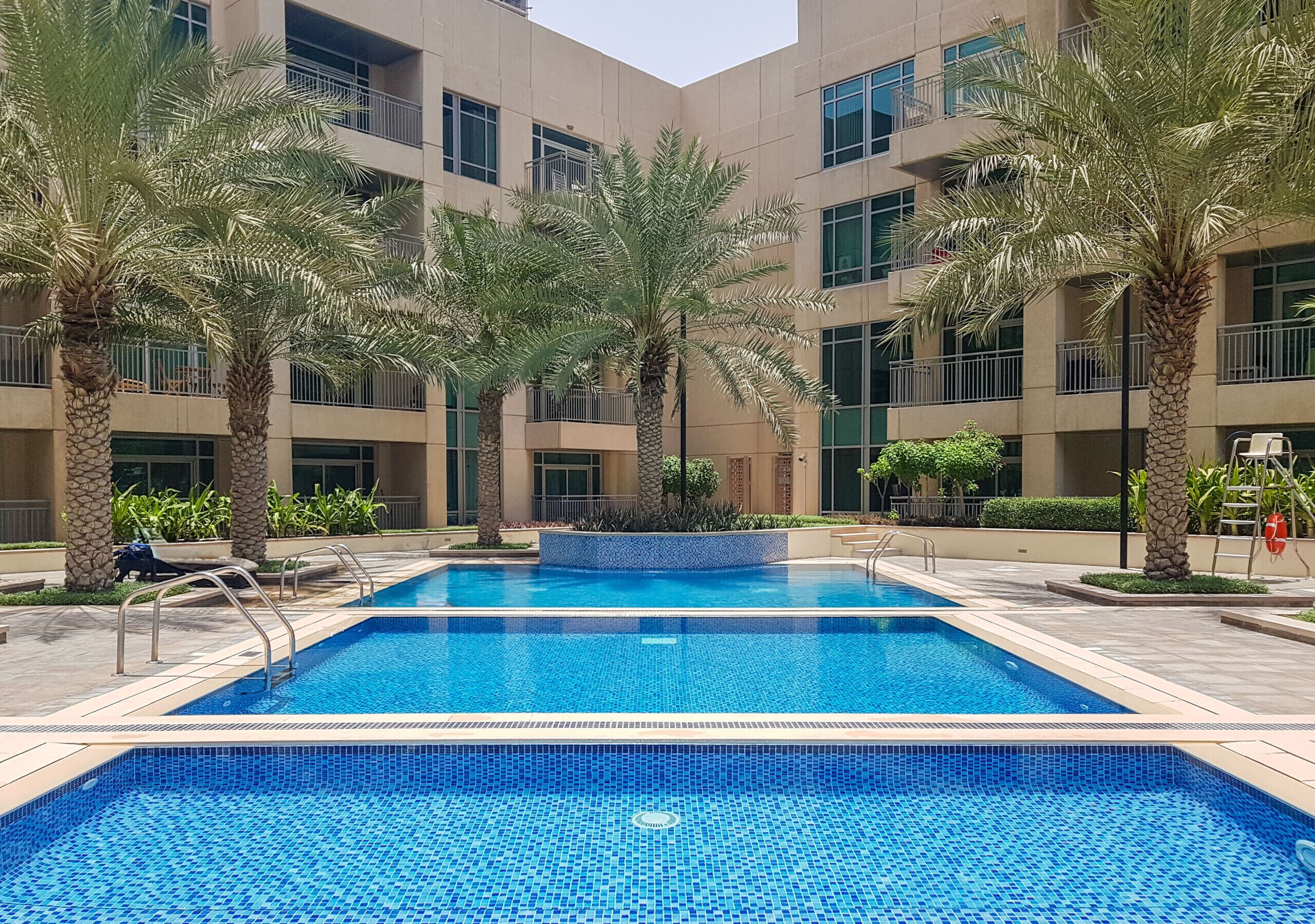 Una piscina con agua cristalina azul, rodeada de palmeras y un edificio de varios pisos con balcones.