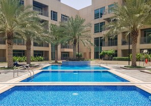 A swimming pool with clear blue water, surrounded by palm trees and a multi-story building with balconies.