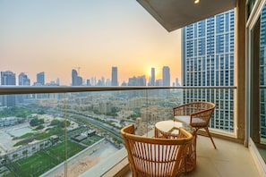 Un balcón con sillas de mimbre y una mesa, con vista a un paisaje urbano que incluye edificios y un campo deportivo.