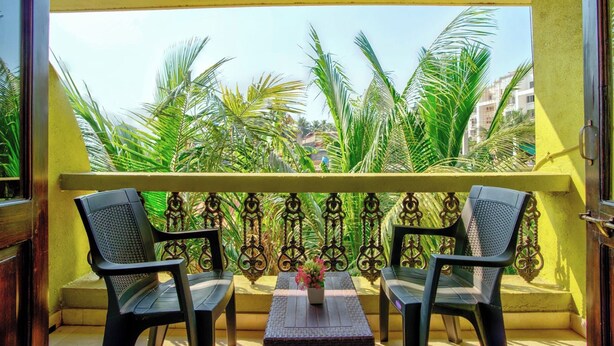 A balcony with two chairs and a table, surrounded by a yellow wall and metal railing, overlooking a view of palm trees and buildings. - Ocean Dream Luxury Villa (Arpora)