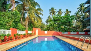 A swimming pool with a white fence, palm trees, and a red building in the background. - Ocean Dream Luxury Villa (Arpora)