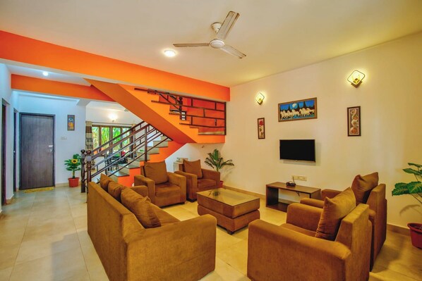 A living room with brown sofas, a coffee table, a TV, and a staircase with orange accents.