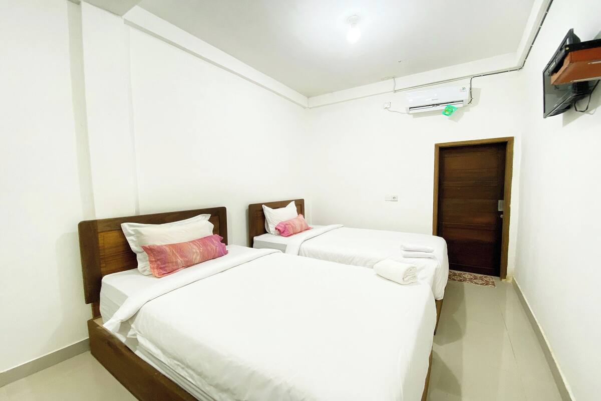 Habitación de hotel con dos camas, ropa de cama blanca, cabecero de madera, una mesita de noche y una puerta.