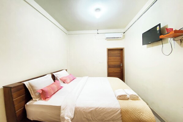 Una cama bien tendida con ropa de cama blanca y rosa, un cabecero de madera y un televisor montado en la pared.