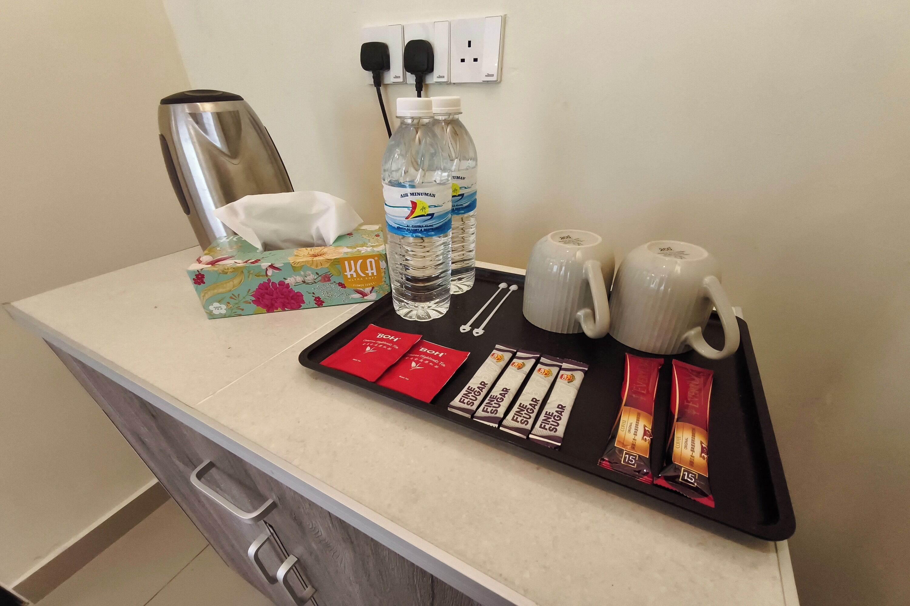 Balcão de quarto de hotel com cafeteira, duas garrafas de água, caixa de lenços, bandeja com sachês de açúcar e duas canecas.