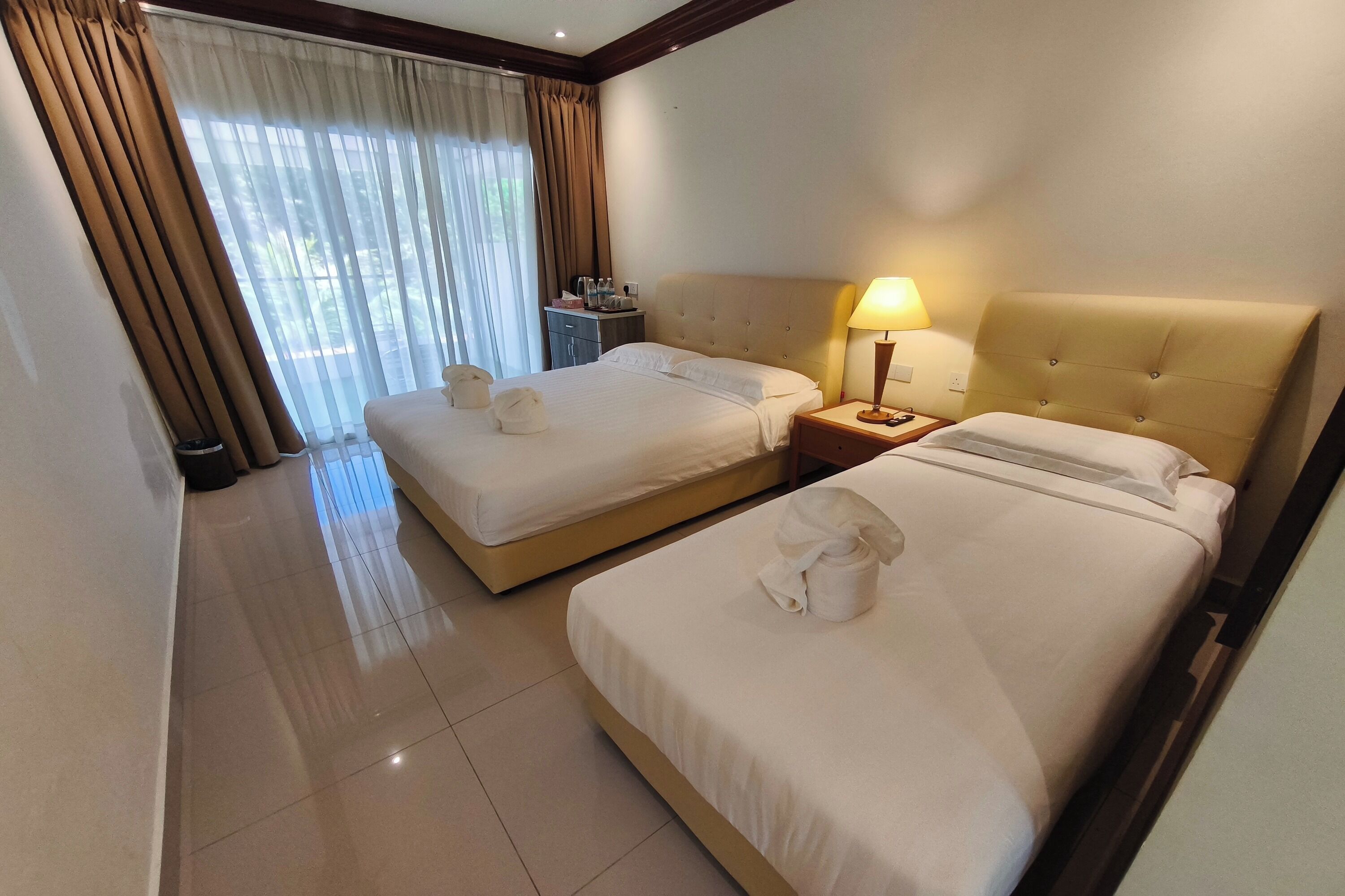 A hotel room with two beds, a bedside table with a lamp, a small table with toiletries, and a window with curtains.