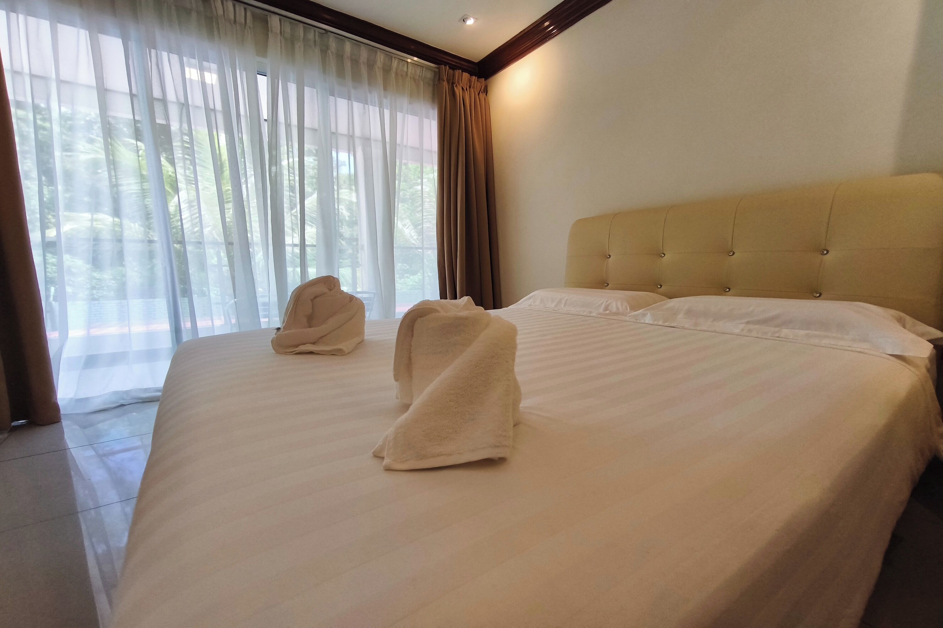 A hotel room with a large bed, two towels on the bed, a headboard, and a view through sheer curtains.